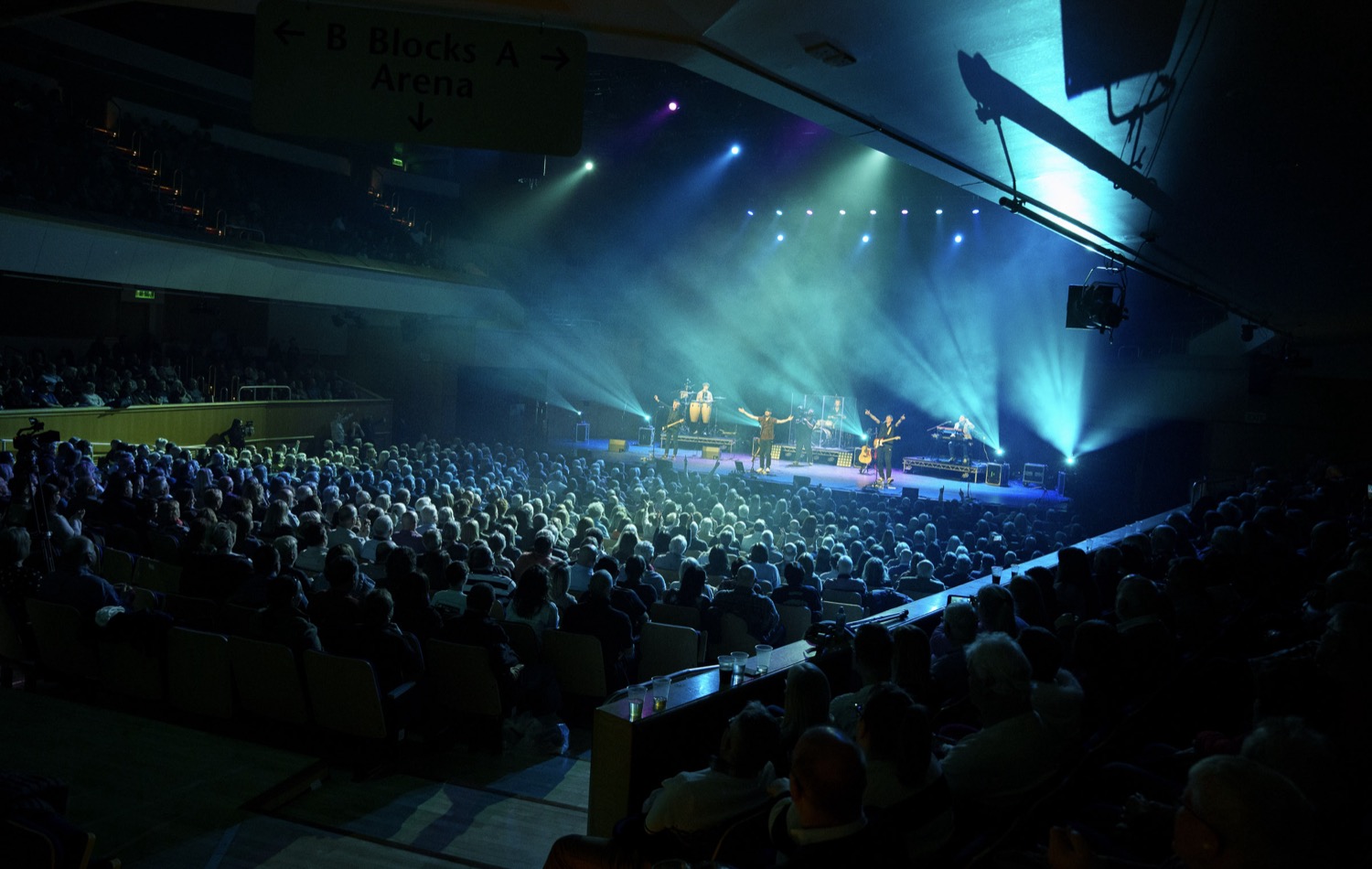 Crowded indoor venue with audience watching a band performing on stage under blue and green spotlights.