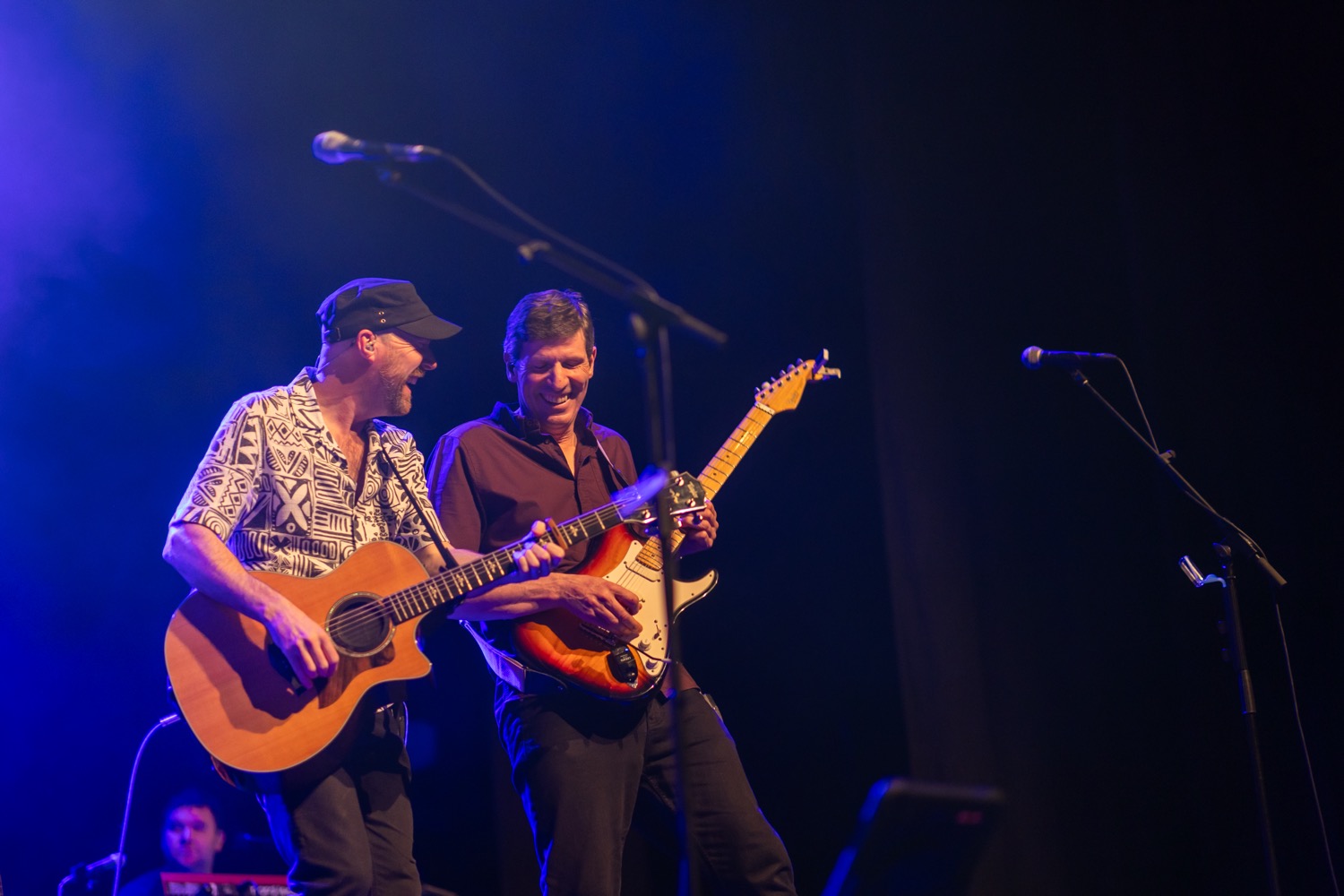 Two male musicians on stage playing guitar and electric guitar, smiling under purple stage lights.