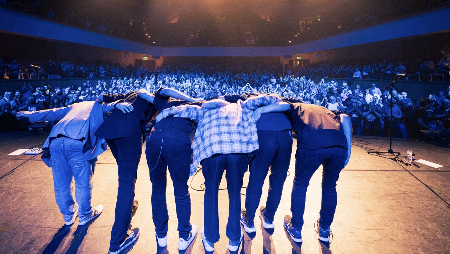 Musicians on stage bowing to an applauding audience in a large, dimly lit concert hall.