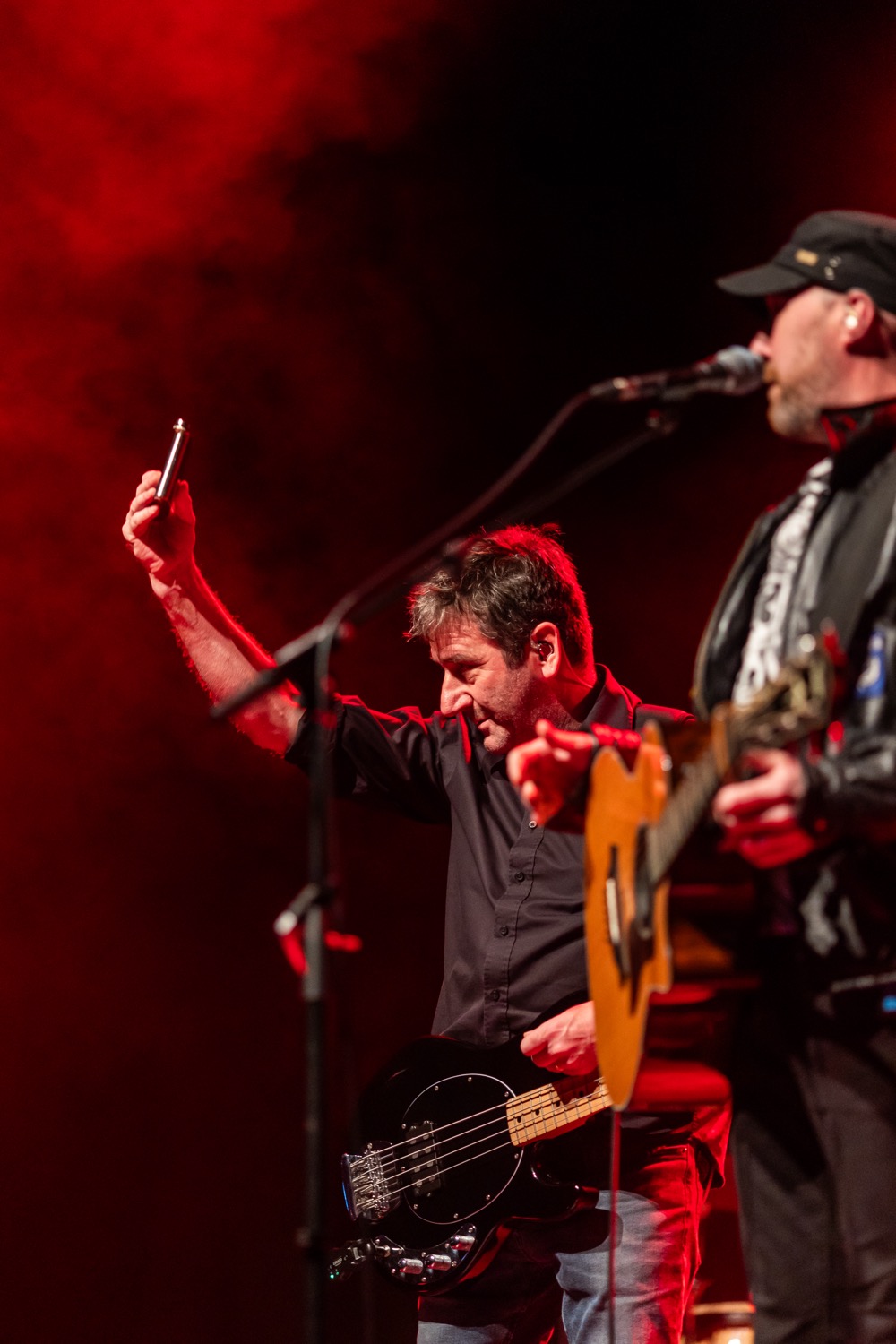 Male musician playing bass guitar holding a harmonica up on stage with red lighting next to a guitarist singing into a microphone.