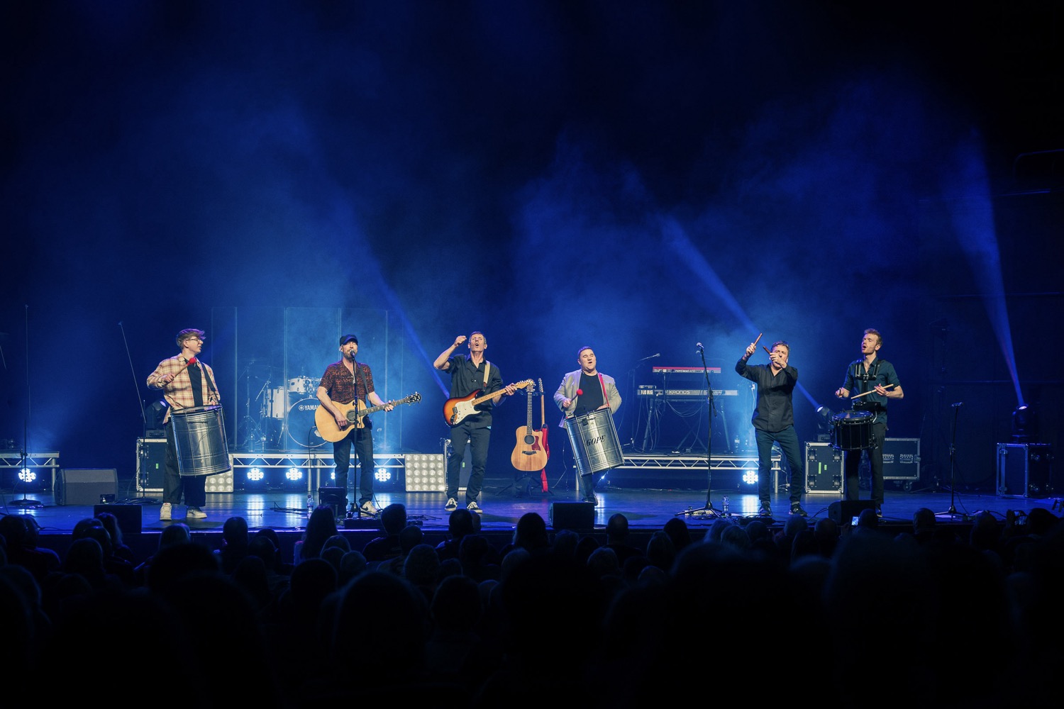 Six musicians performing on stage with guitars and drums under blue stage lights in front of a seated audience.