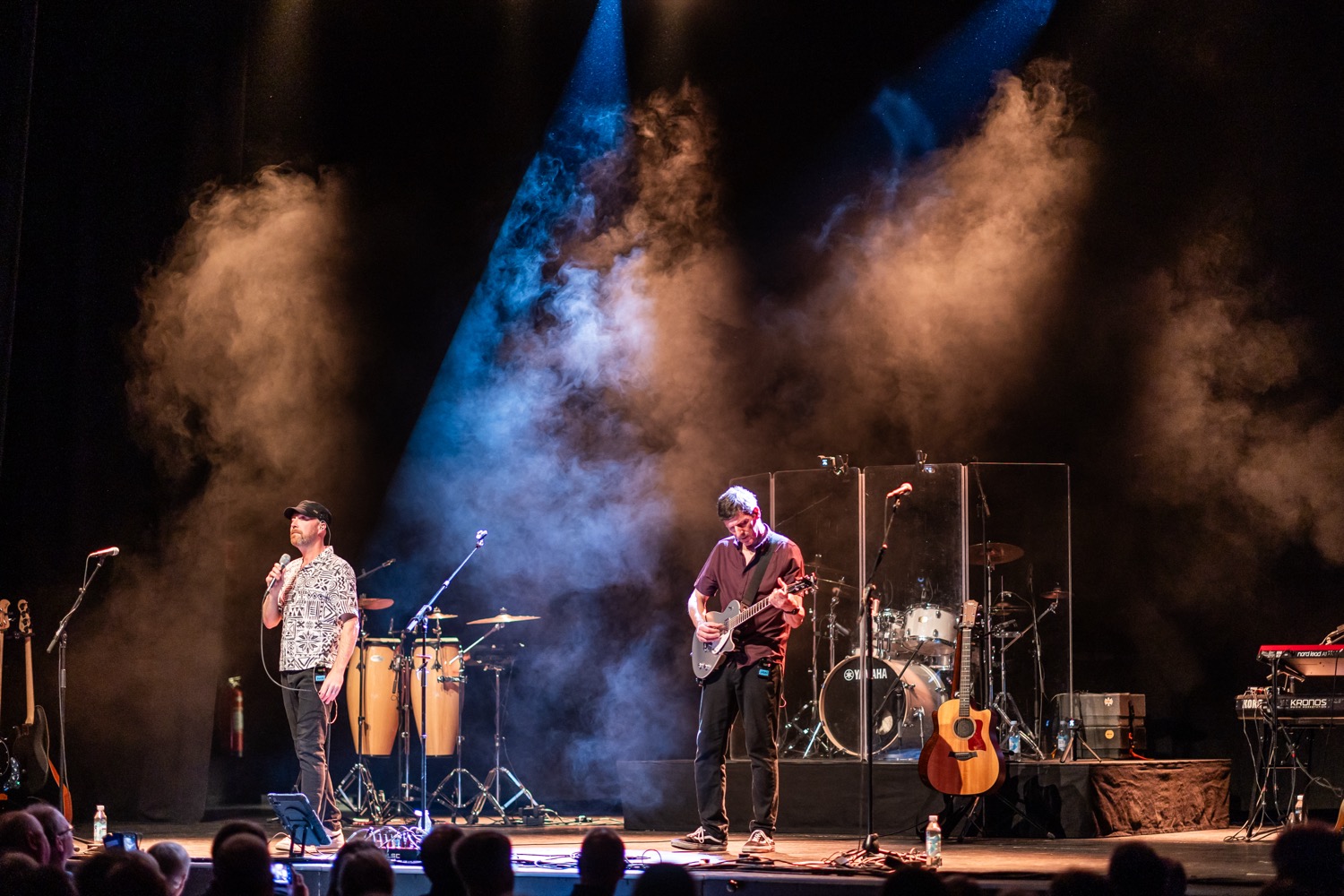 Two musicians on stage with one singing into a microphone and the other playing an electric guitar under blue and orange stage lights with smoke effects.