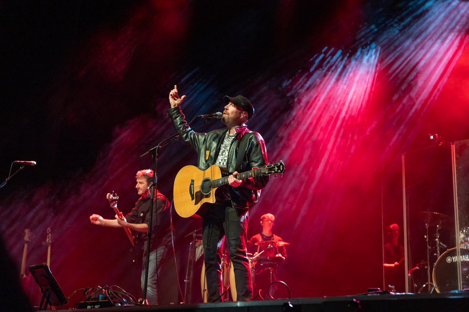 Musician in a black leather jacket and cap plays acoustic guitar and sings on stage with band members under red and blue stage lights.