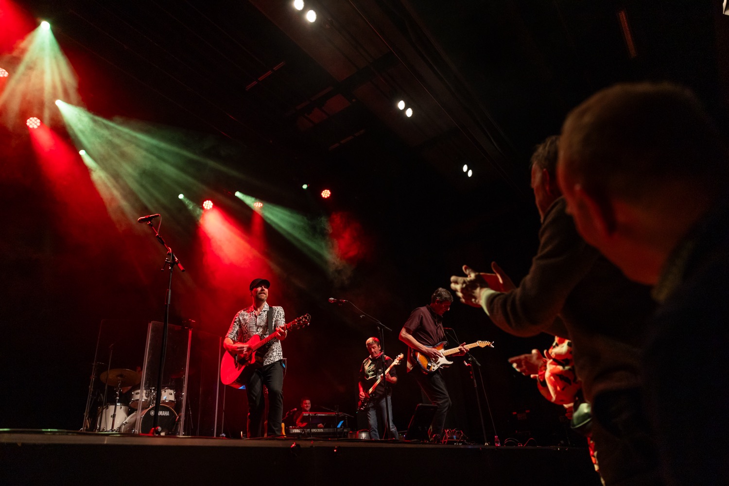 Band performing on stage with red and green spotlights and an audience clapping in the foreground.
