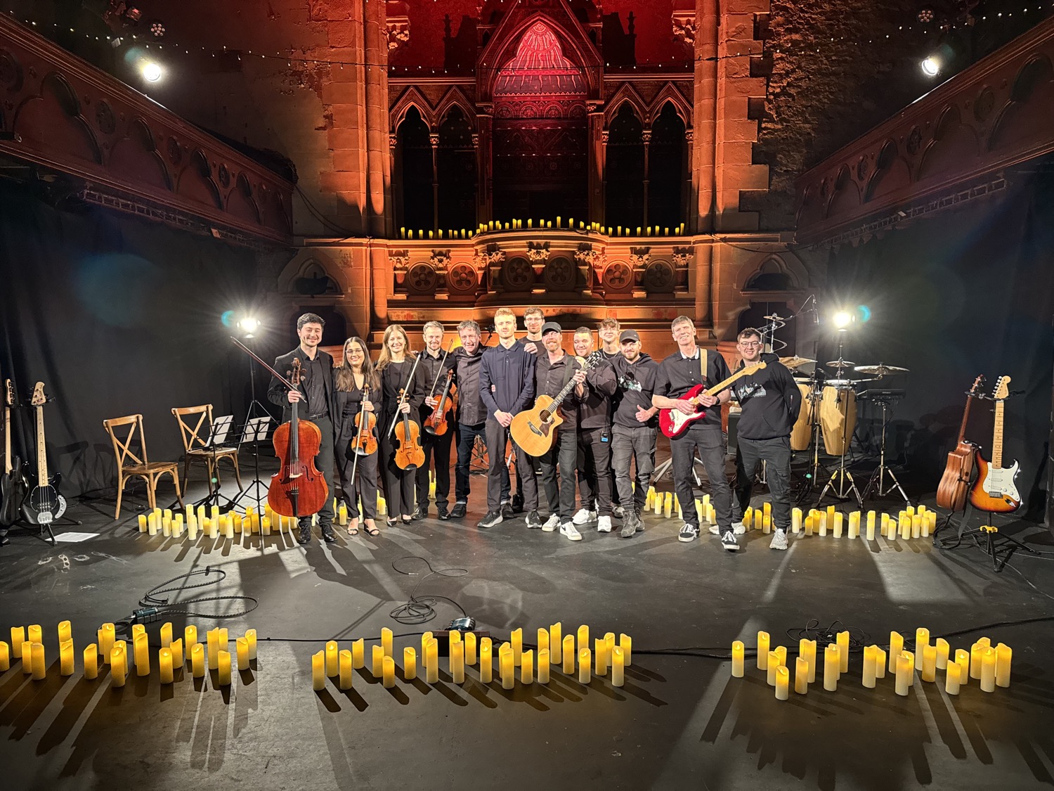 Group of musicians standing on stage in a dimly lit venue with lit candles arranged on the floor around them, holding various string and electric instruments.