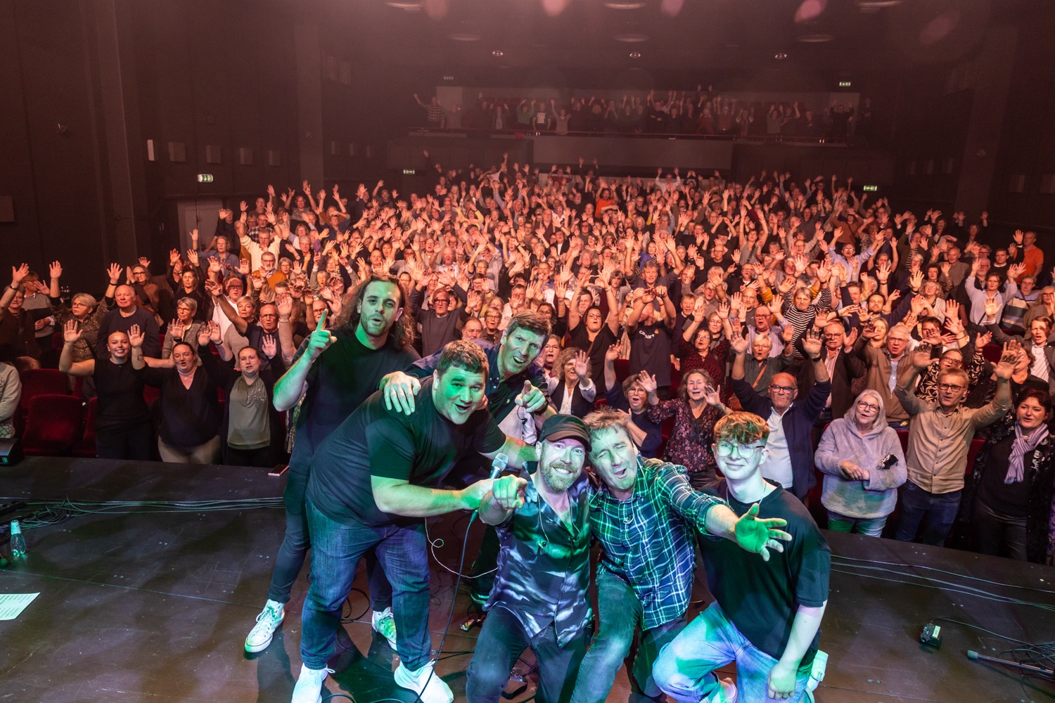 Six men on a stage posing for a selfie with a large crowd behind them, all smiling and waving.