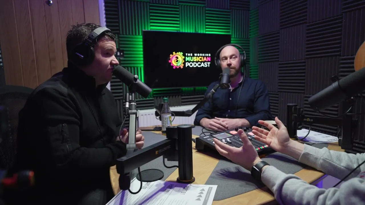 Three men wearing headphones and speaking into microphones in a soundproof podcast studio with a screen displaying The Working Musician Podcast logo.