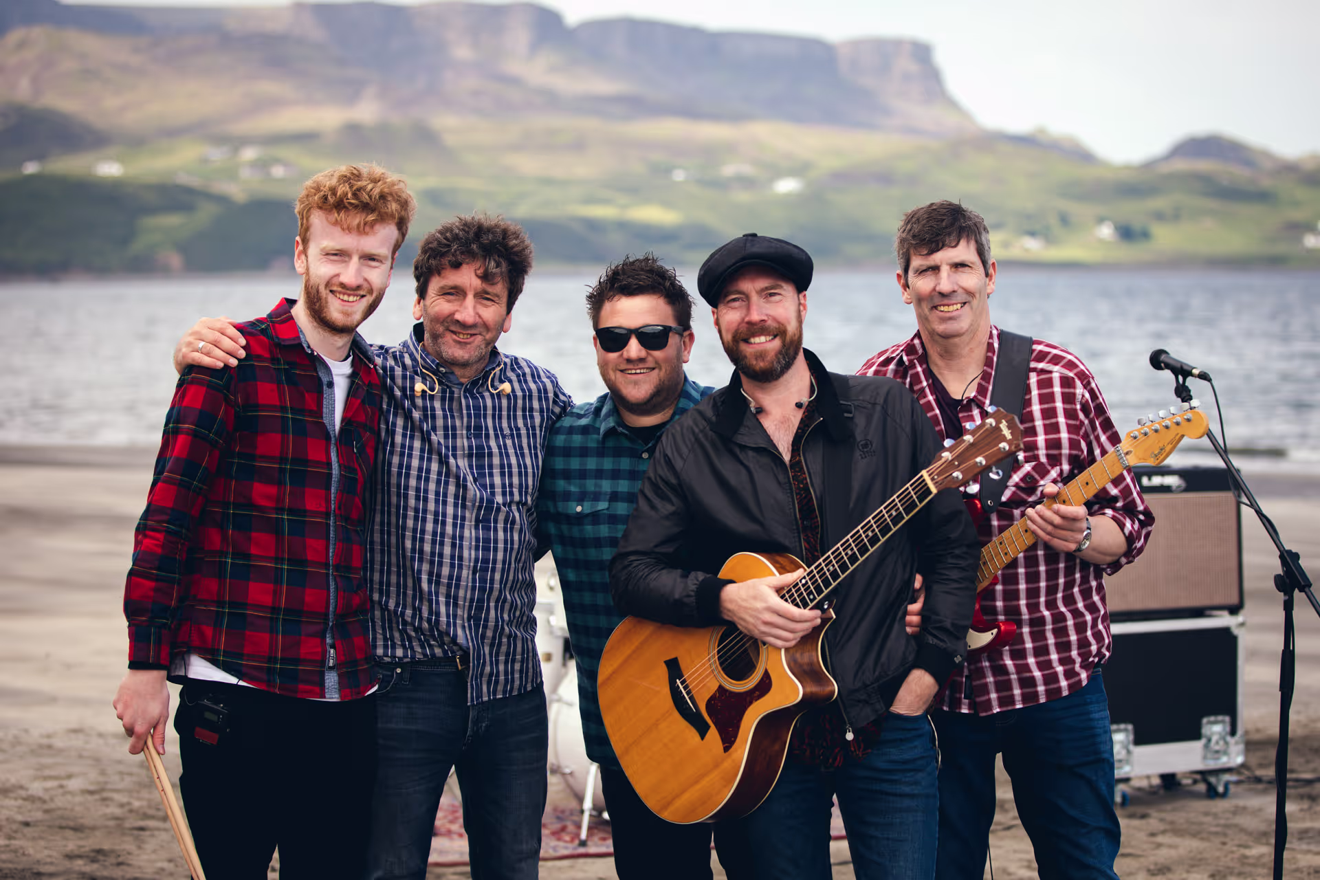 Five men posing with musical instruments on a beach with mountains and water in the background.