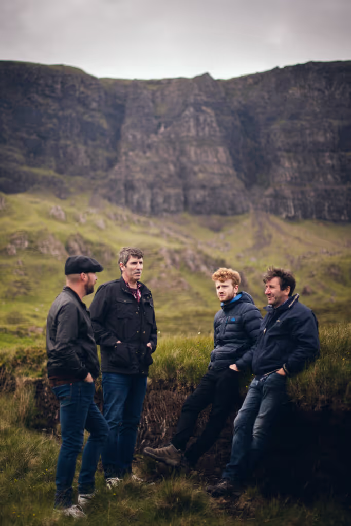 Four men in jackets standing and leaning on grass with rocky cliffs in the background.