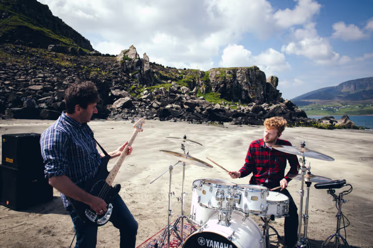 Two musicians playing bass guitar and drums on a sandy beach with rocky hills and a cloudy sky in the background.