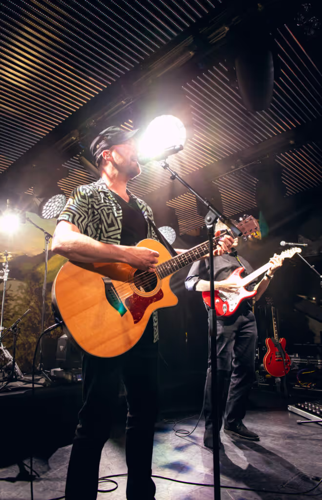 Two musicians playing guitars on stage under bright spotlights, one with an acoustic guitar and the other with an electric guitar.