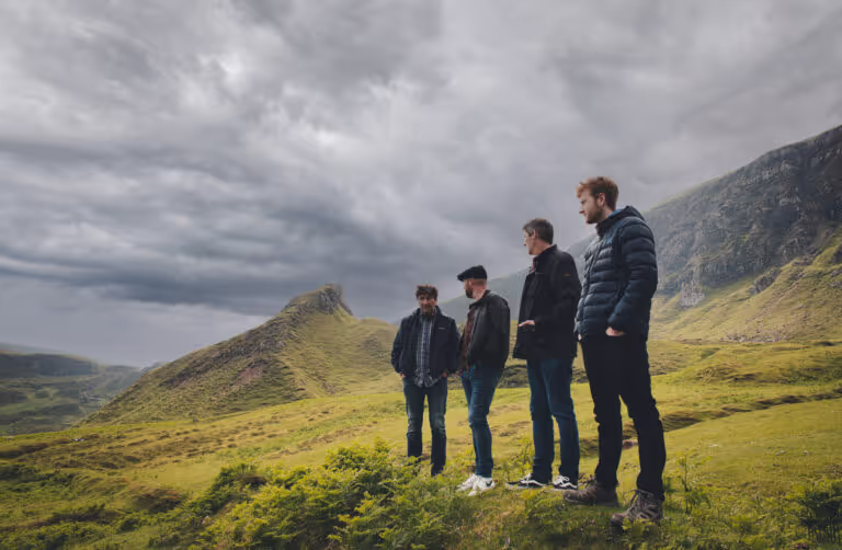 Four men standing on a grassy hillside with cloudy sky and rocky mountains in the background.