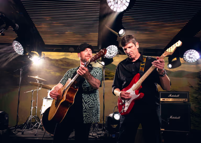 Two male musicians performing on stage, one playing an acoustic guitar and the other playing a red electric guitar under bright stage lights.