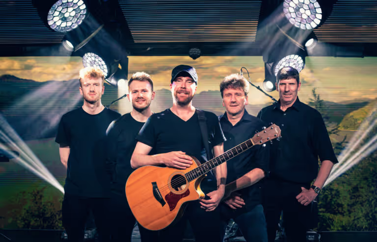 Group of five men on stage with one holding an acoustic guitar, illuminated by stage lights.