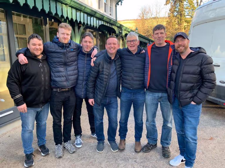Seven men dressed in casual winter jackets and jeans standing arm-in-arm outside near a green building and white van.