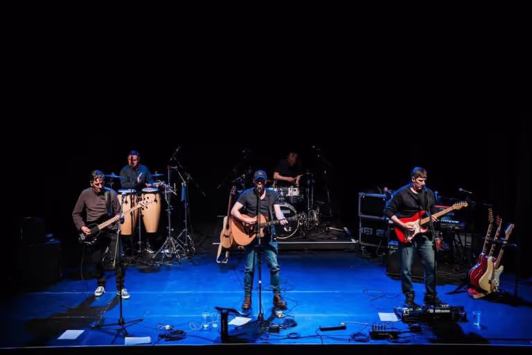 Five-member band performing on stage with instruments including electric guitars, acoustic guitar, drums, and congas under spotlights.