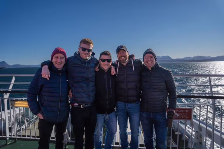 Five men dressed in winter jackets and hats standing close together, smiling on a boat deck with a sunny ocean and mountainous background.