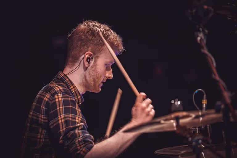 Young man playing drums with drumsticks in hand against a dark background.