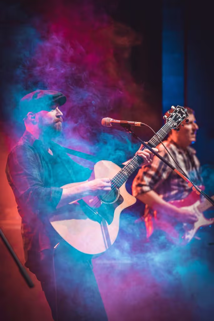 Two musicians playing guitars on stage surrounded by colorful blue and pink smoke.