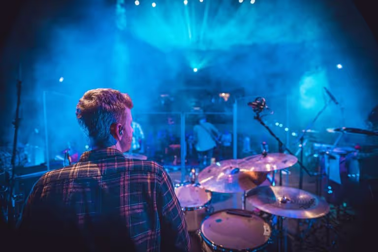 Drummer playing on a drum set during a live concert with blue stage lights and smoke.