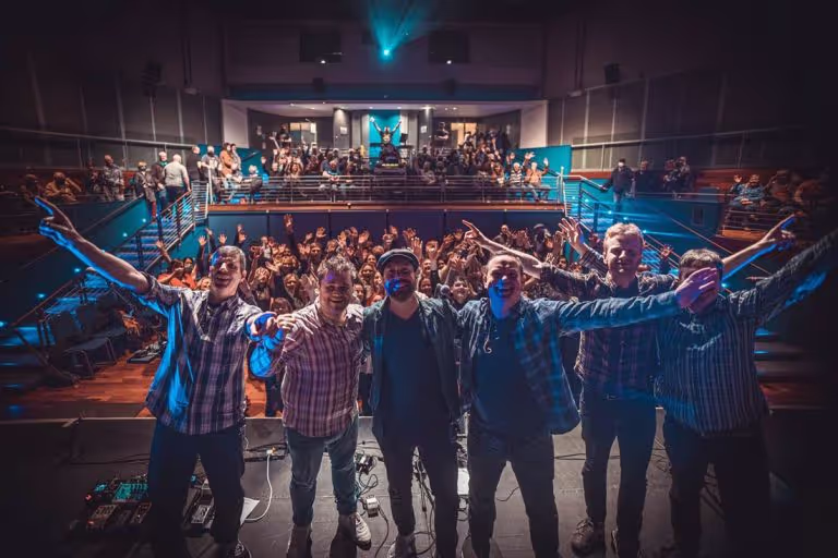 Six men standing on a stage in front of a cheering audience in a theater with their arms raised.