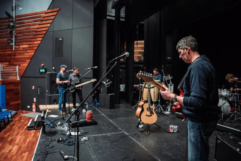 Musicians rehearsing with guitars, drums, and microphones on a stage with wooden panels and black curtains.