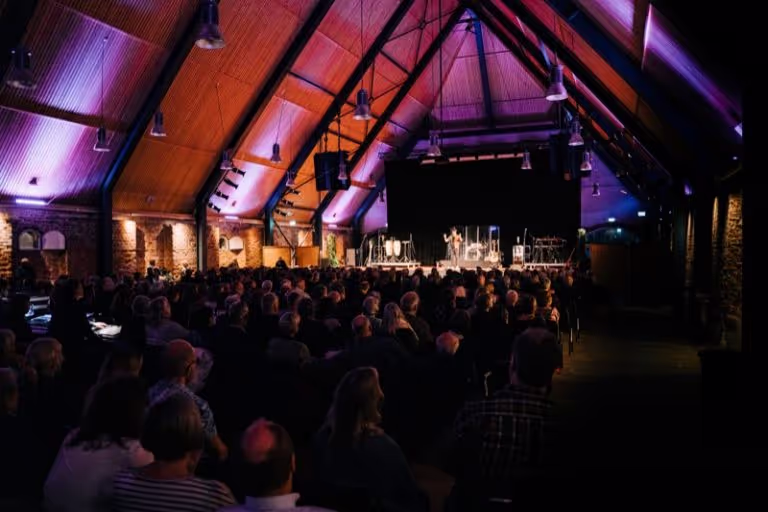 Audience seated in a large hall with wooden vaulted ceiling watching a person speaking on stage under purple lighting.