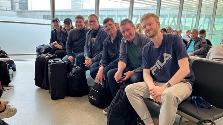 Seven men sitting closely on airport seats with luggage, smiling and posing for the photo.