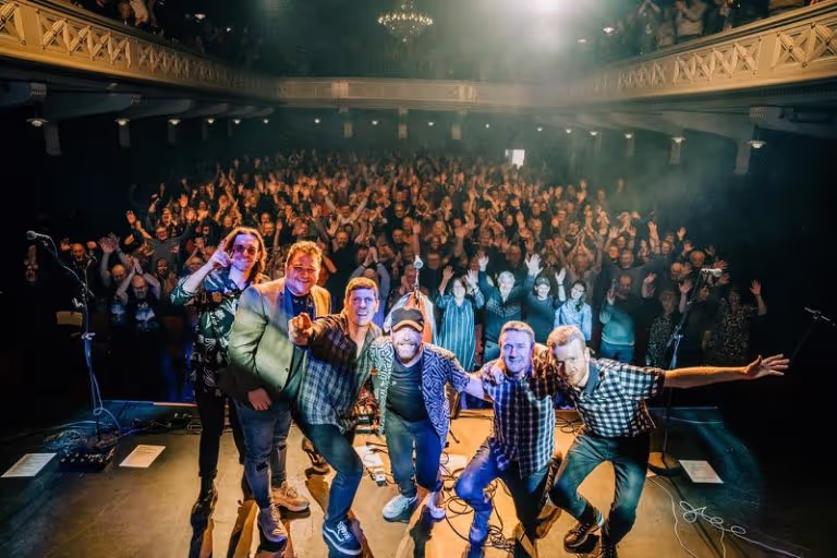 Five band members posing on stage with a large cheering audience behind them in a theater.