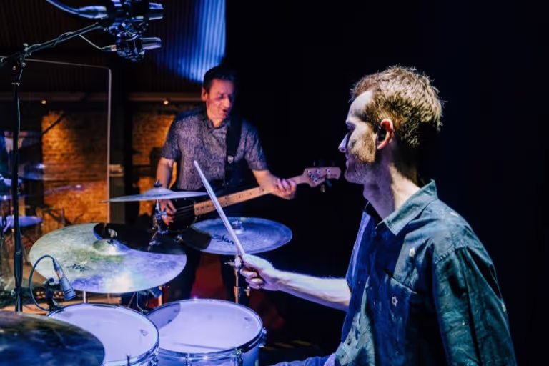 Man playing drums with drumsticks while another man plays bass guitar in a dimly lit room.