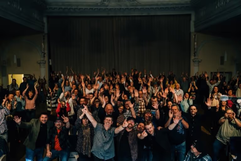 Large group of people in a dimly lit room smiling and waving towards the camera.