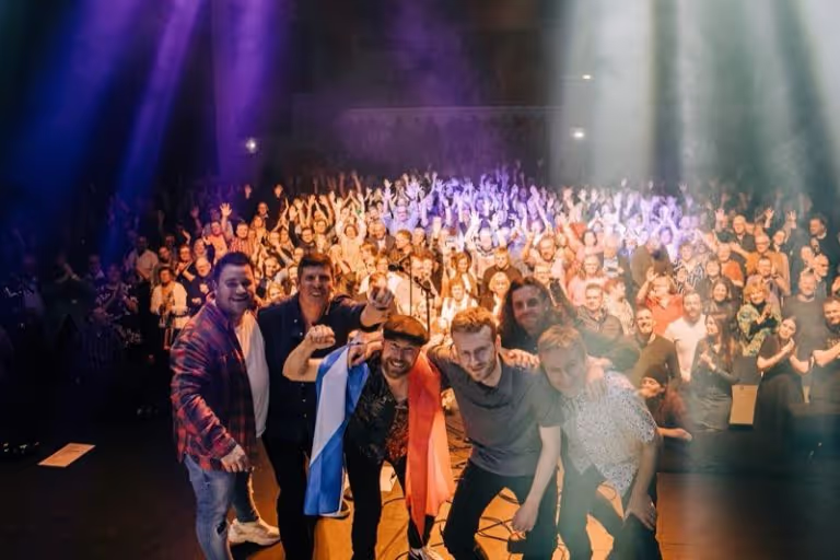 A band of six men posing happily on stage in front of a cheering crowd with colorful stage lights shining.