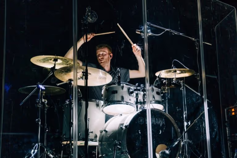 Drummer playing a Yamaha drum set behind a transparent sound shield on a dark stage.
