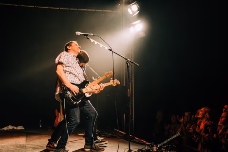 Two musicians playing electric guitars on a stage under warm spotlights with an audience watching.