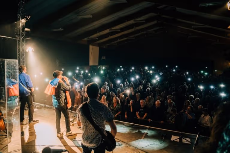 Musicians performing on stage with an audience holding up lit smartphones in a dark venue.