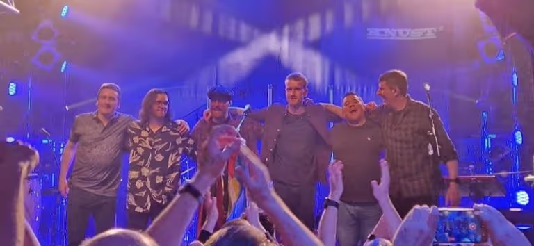 Six musicians standing arm-in-arm on stage under blue and purple lights, with audience members clapping and taking photos in the foreground.
