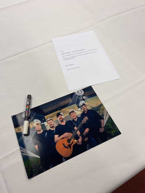 A photograph of a five-member band with one holding an acoustic guitar, placed on a white surface next to a black marker and a printed note.