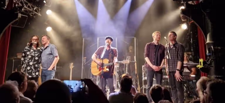 Five musicians performing on a stage with spotlights and a crowd watching, one playing acoustic guitar and two singing into microphones.