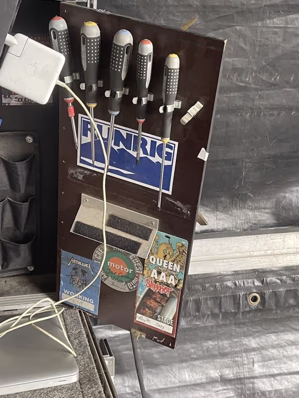 A wooden board with five screwdrivers attached, backstage passes for Queen and Slipknot, and an Apple power adapter with cable.