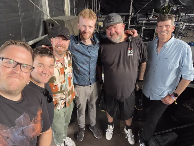 Group of six men standing closely together smiling backstage at an event.