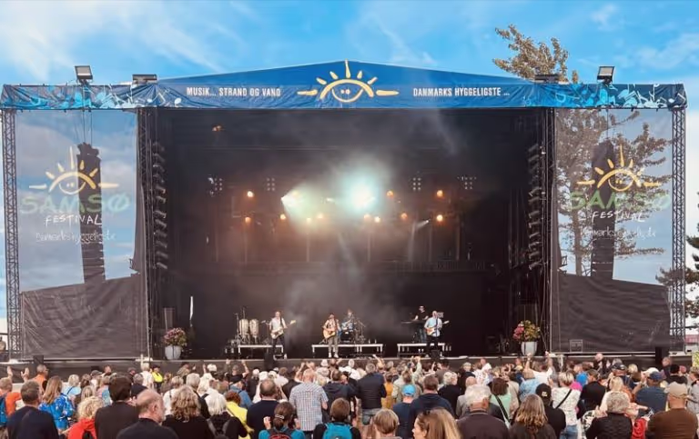 Outdoor music festival stage with a live band performing under bright lights and a crowd watching.