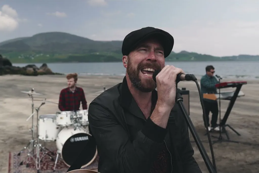 Man wearing a black cap singing into a microphone with a drummer and keyboard player behind him on a beach with mountains in the background.