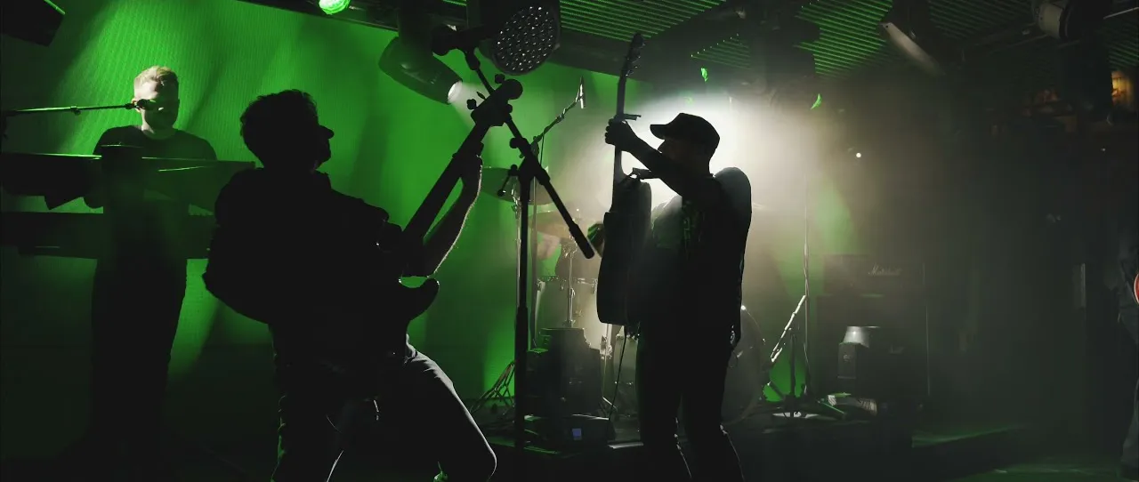 Silhouettes of band members playing guitar and keyboard on stage with green backlighting and smoke effects.