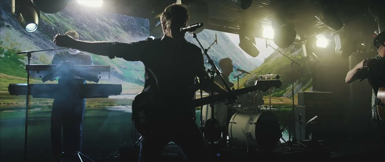 Silhouetted band performing on stage with keyboardist, guitarist, and drummer in front of a mountainous landscape backdrop.