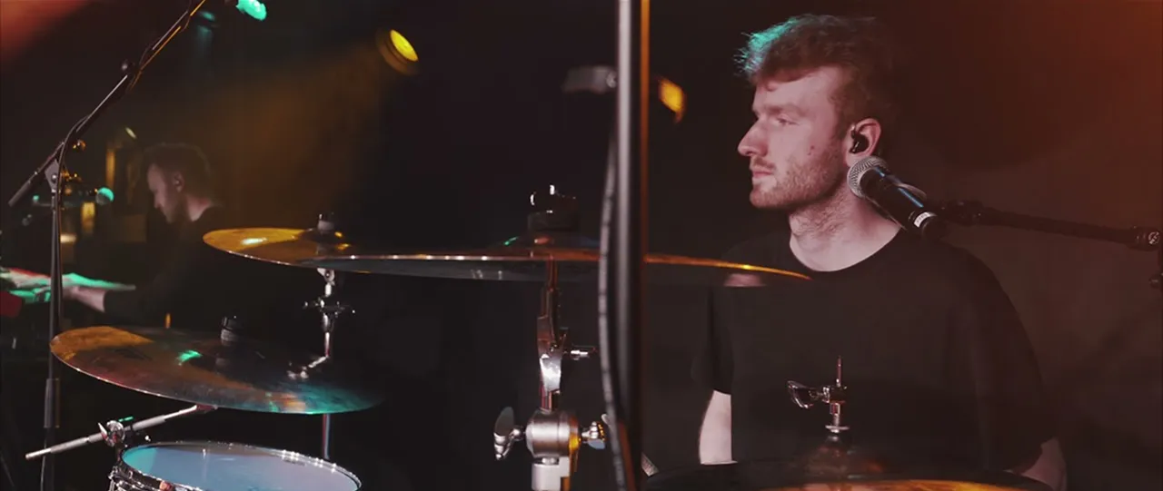 Side view of a drummer wearing a black shirt sitting behind a drum set with cymbals and a microphone, with a keyboard player blurred in the background in a dimly lit stage setting.