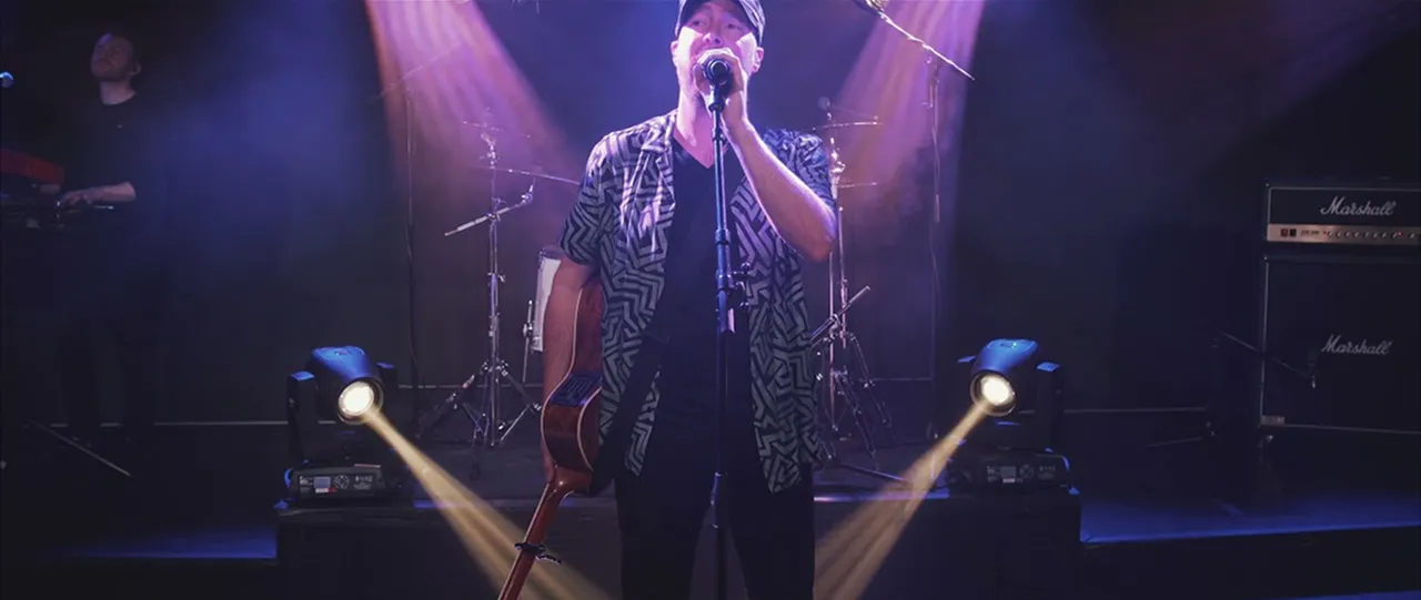 Male singer holding a microphone and guitar on stage with drum set and keyboard player in background, illuminated by purple spotlights.