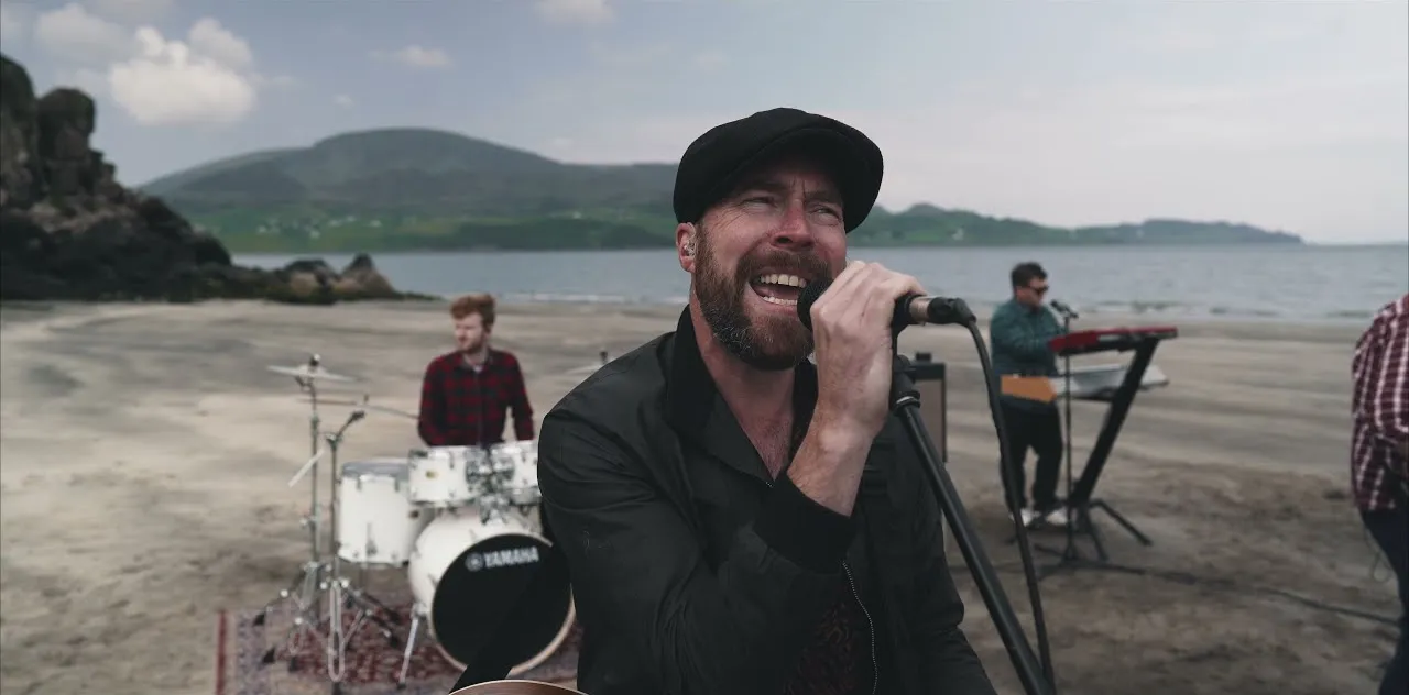 Bearded man in black jacket and cap singing into microphone on a beach with band members playing drums and keyboard in the background.